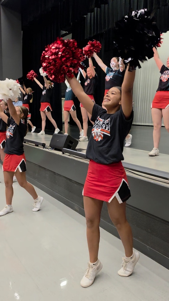 Group of cheerleaders on a stage with red and black pom-poms, wearing black and red uniforms, performing in front of a black curtain.