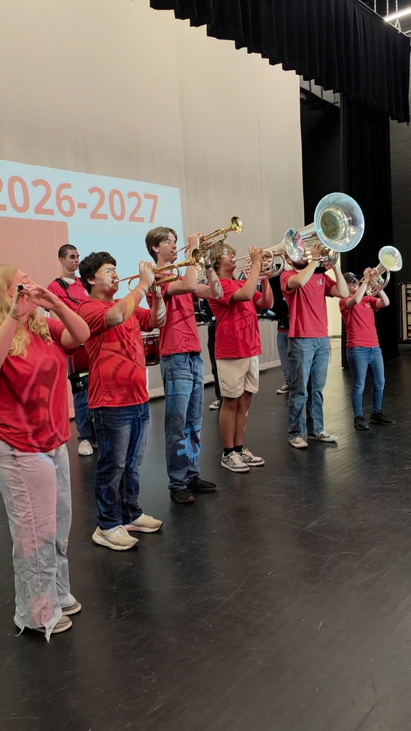A group of musicians wearing red shirts and playing instruments on a stage with a 2026-2027 backdrop.
