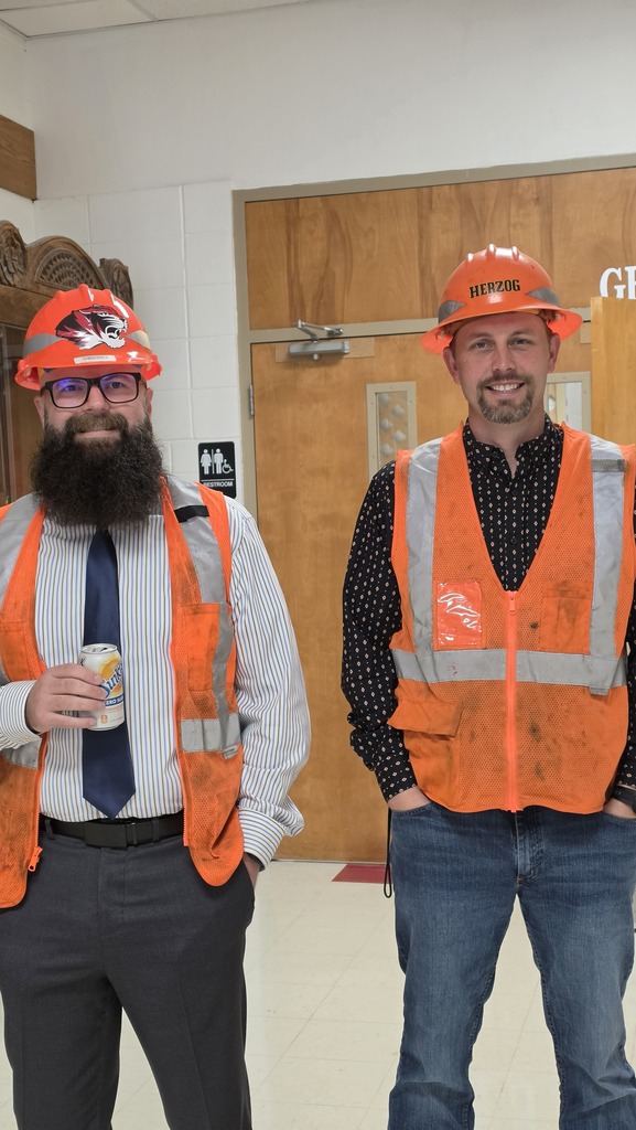 Two men stand together wearing hard hats and construction vests. 