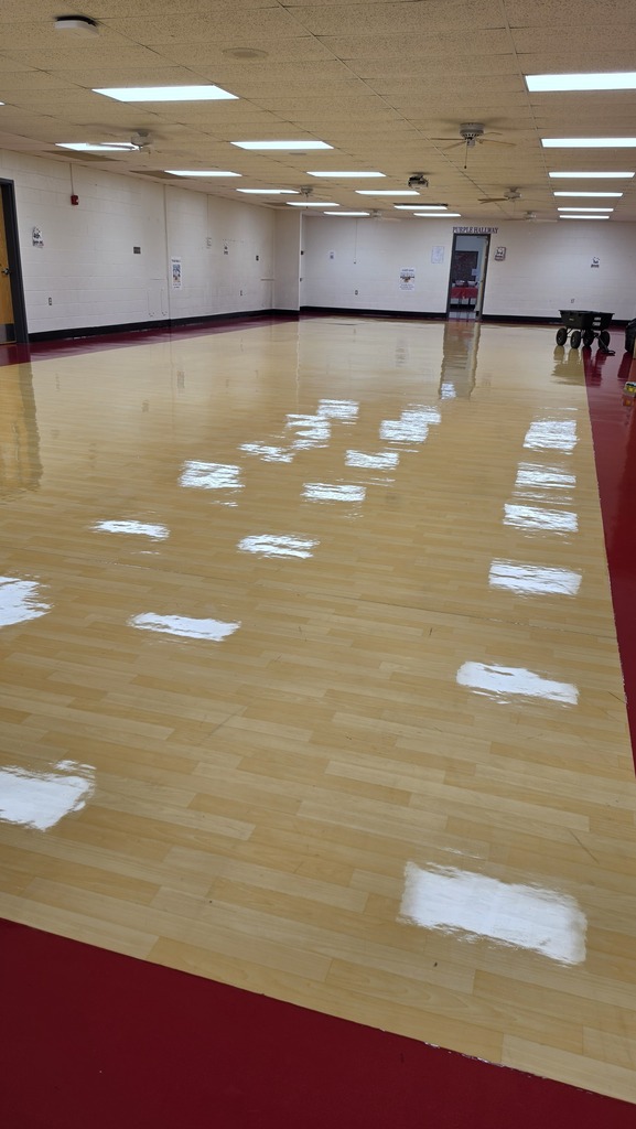 A vast room with white walls and ceiling lights. A polished wooden floor and red floor trim.