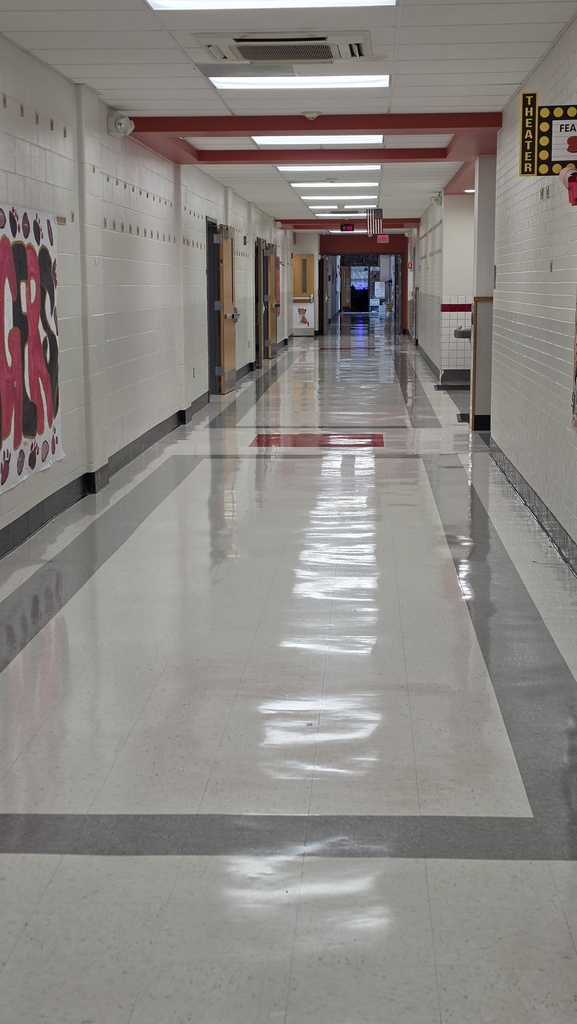 An empty school hallway with white tiles, red and gray stripes, and a row of fluorescent lights along the ceiling.