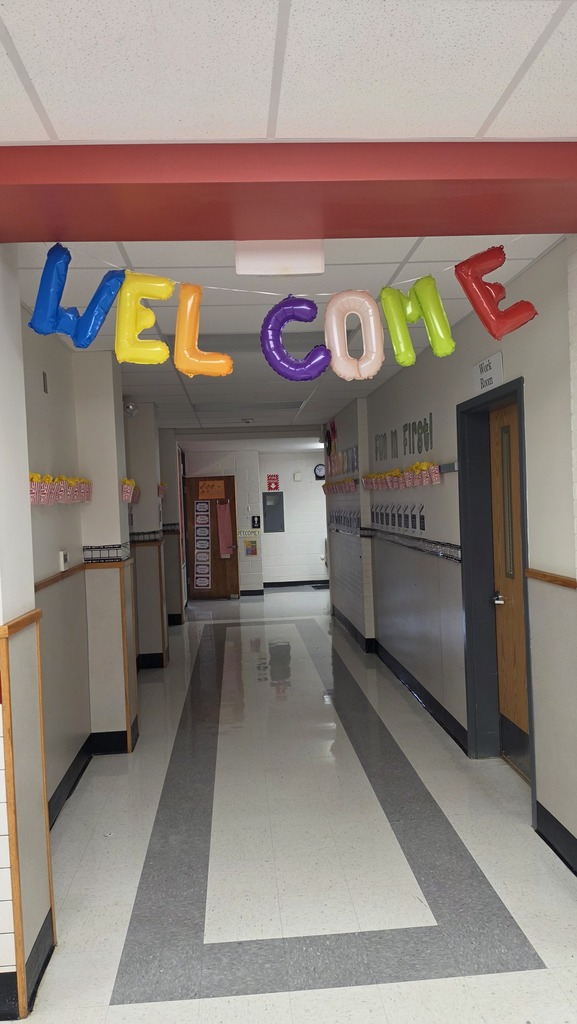 An empty hallway with a colorful "Welcome" banner above and white and gray flooring. Walls are white with red trim.