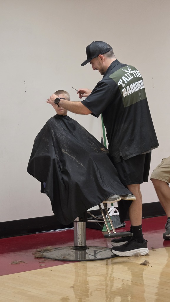 Man in black hat and shirt trims hair of a seated person in a barber's chair.