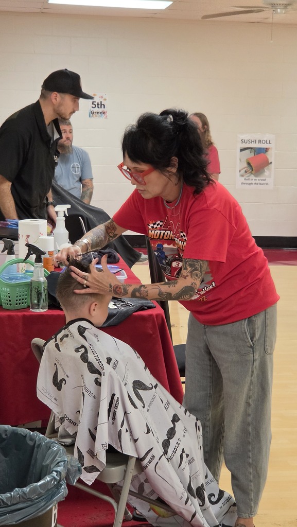 A woman in red shirt and gray pants cuts a boy's hair in a chair at a table.