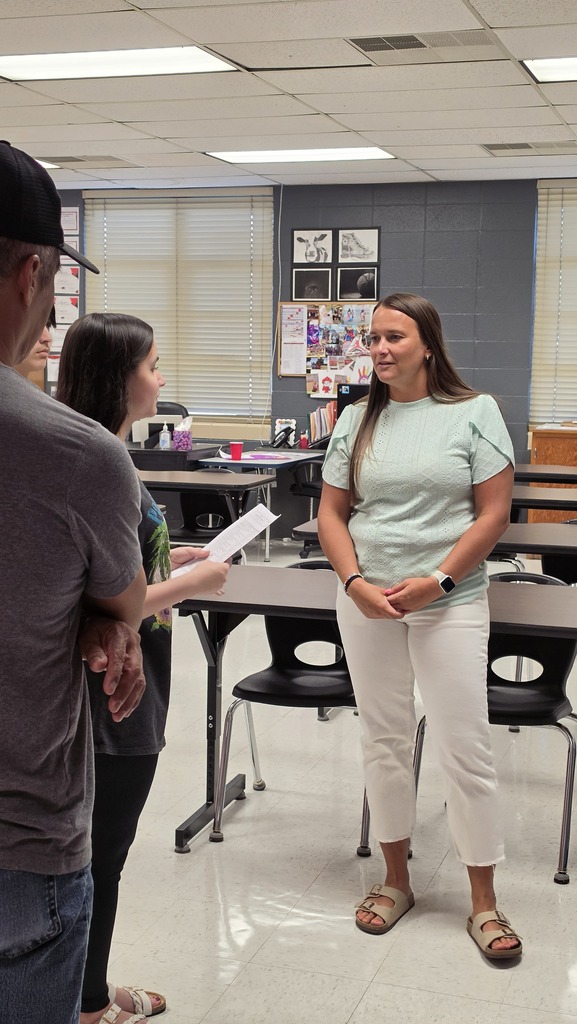 Three people are standing in a classroom. A woman holds a piece of paper and speaks.
