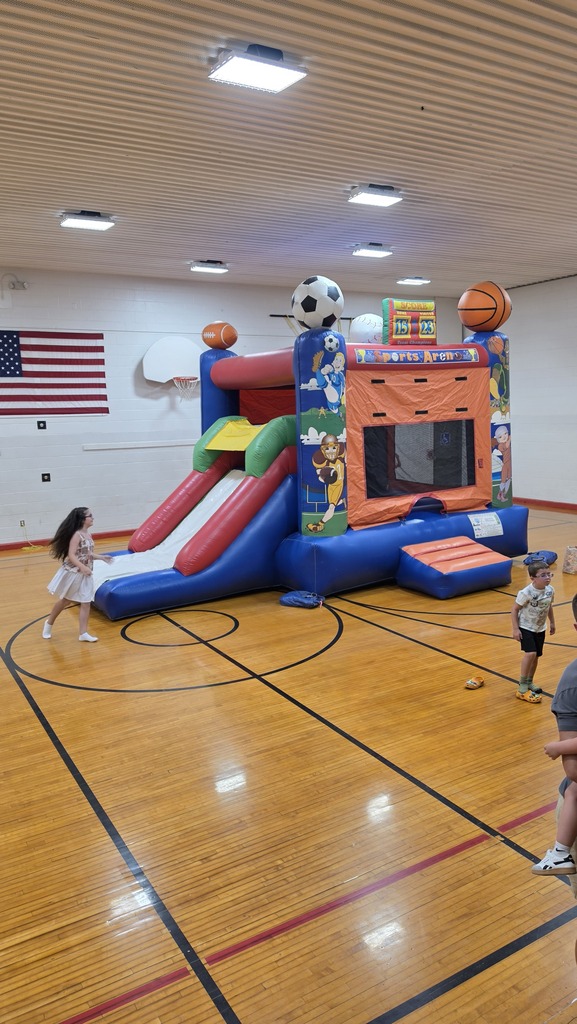 A basketball court with children playing. A colorful inflatable with a slide is in the center. An American flag is in the background.