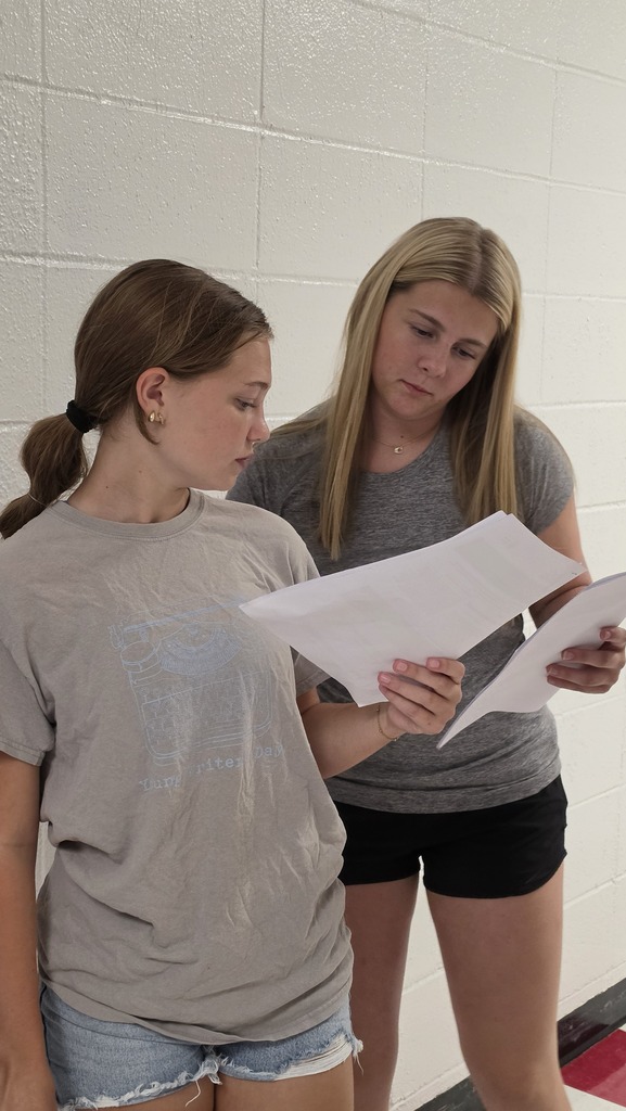 Two females in shorts and tees, one with a ponytail, are reading paper inside.