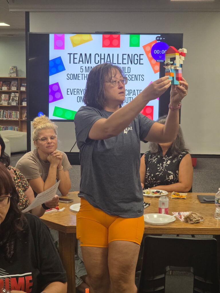 Woman holds a Lego creation while standing at a table in a meeting room with a projector screen behind.