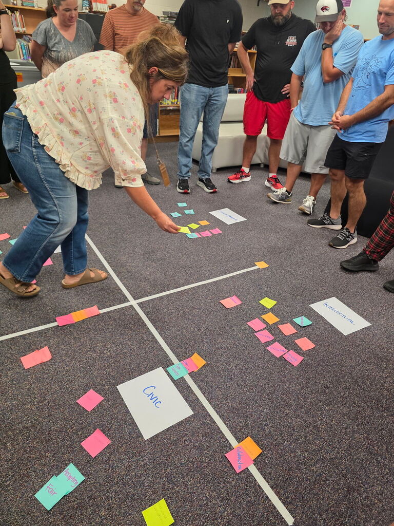 Group of people in a room with a carpet floor. One woman bends over to pick up a sticky note.