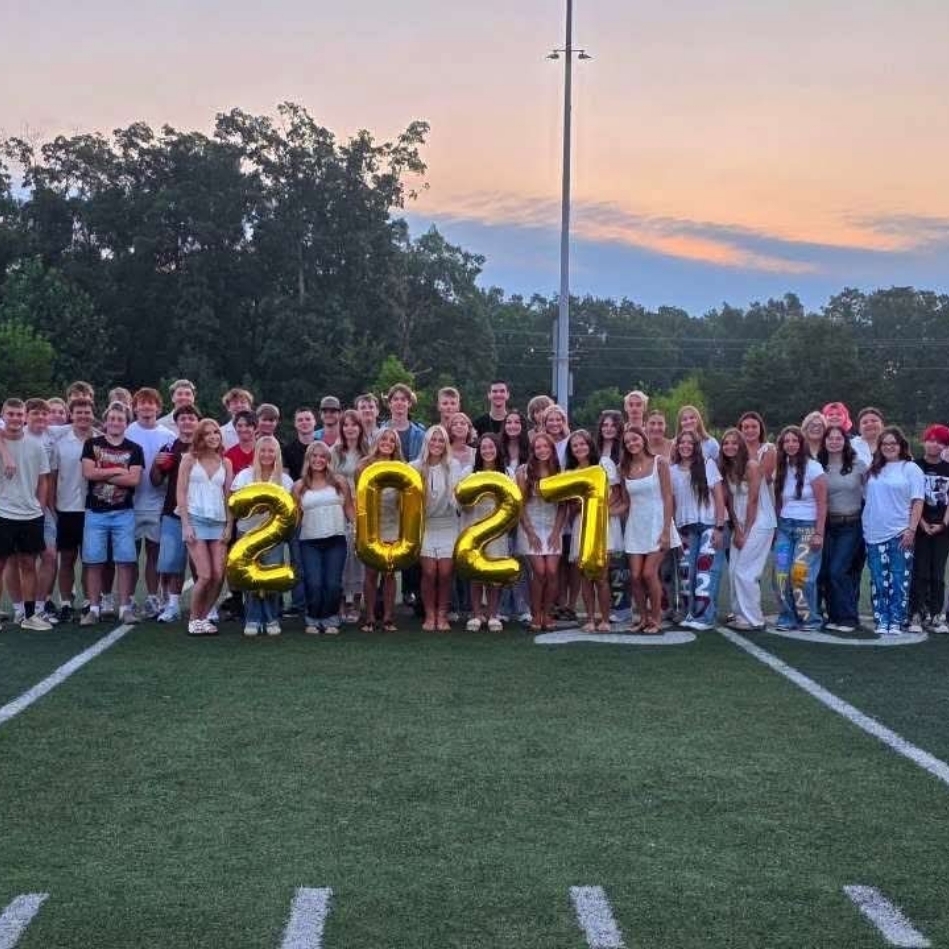 A large group of individuals stand on a football field with white lines and a sunset backdrop.
