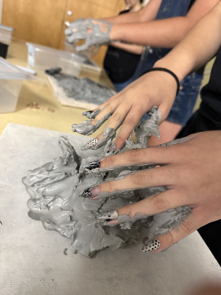 A person's hands with gray clay on them, molding it into a form on a table.