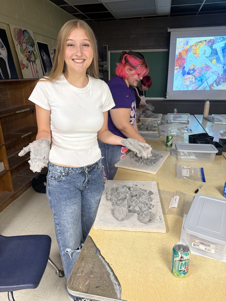 A woman stands at a table with clay, smiling. Another person in pink hair works behind her.