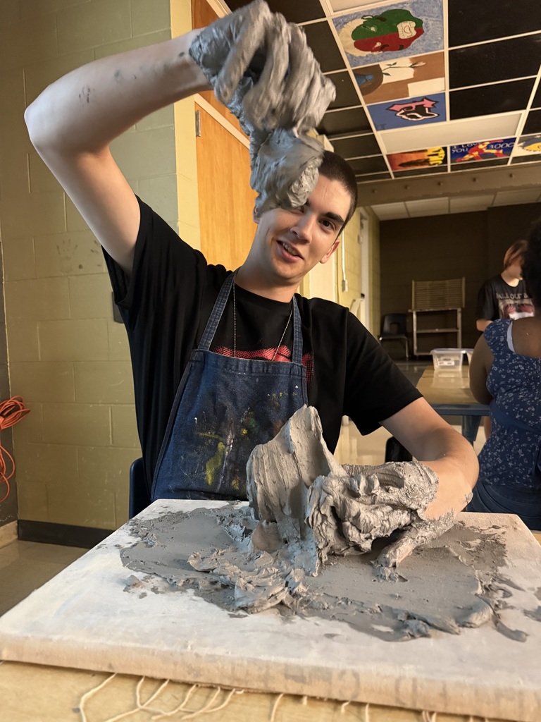 Person wearing an apron works with gray clay on a table, indoors. Behind, another person stands.