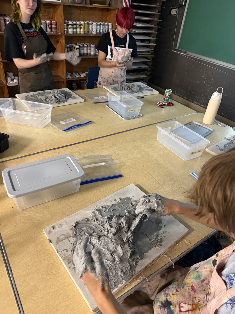 Several individuals work in a studio. Three of them stand behind a table covered in clay, tools, and other materials.