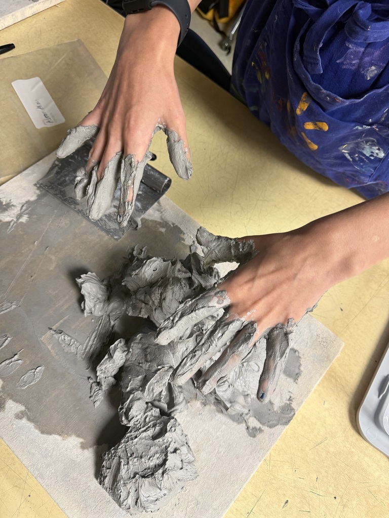 A person wearing a blue apron is molding clay on a table. The clay is gray and wet.