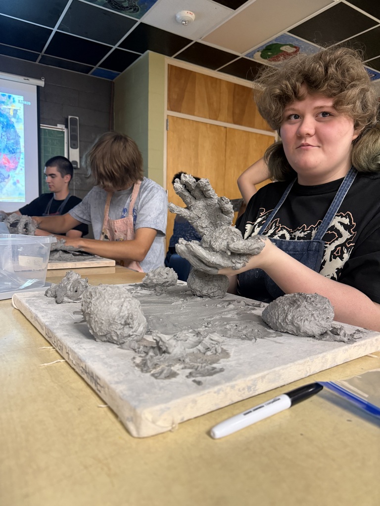 A student with curly hair and wearing an apron molds clay on a table in a classroom.