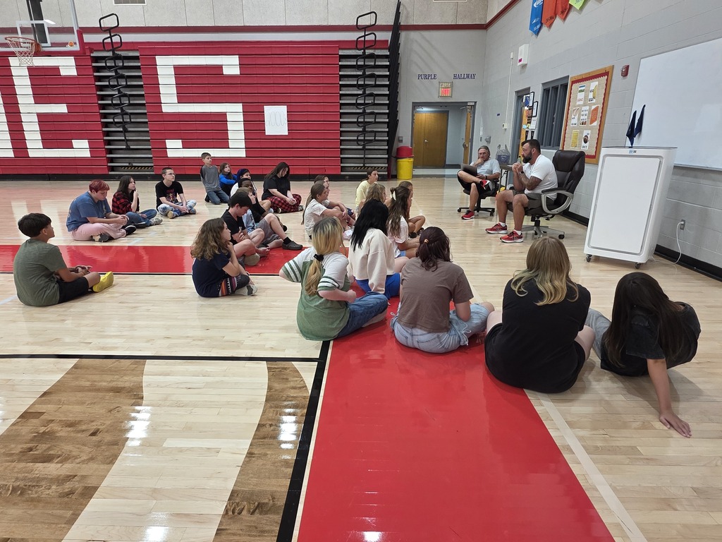 A group of people in a gym, sitting on the floor around a red mat. Some adults are seated on chairs.