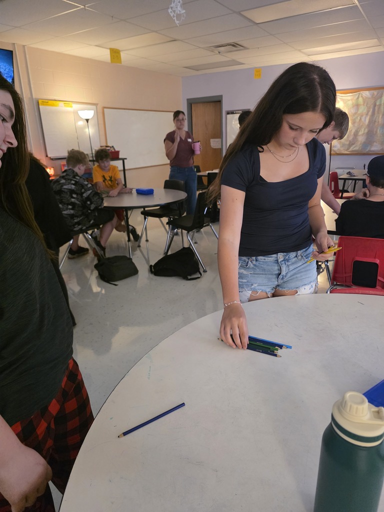 A group of people are gathered around a table in a classroom. One person stands at a table.