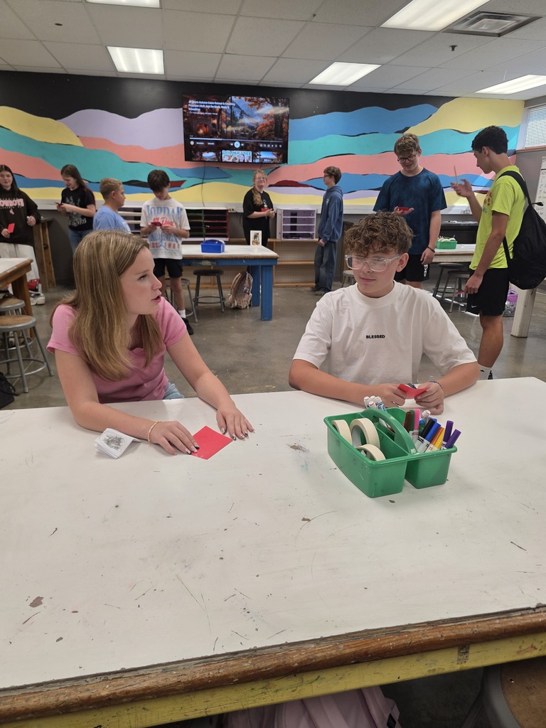 A classroom with students sitting at tables. A boy and girl are sitting at a table with art supplies.