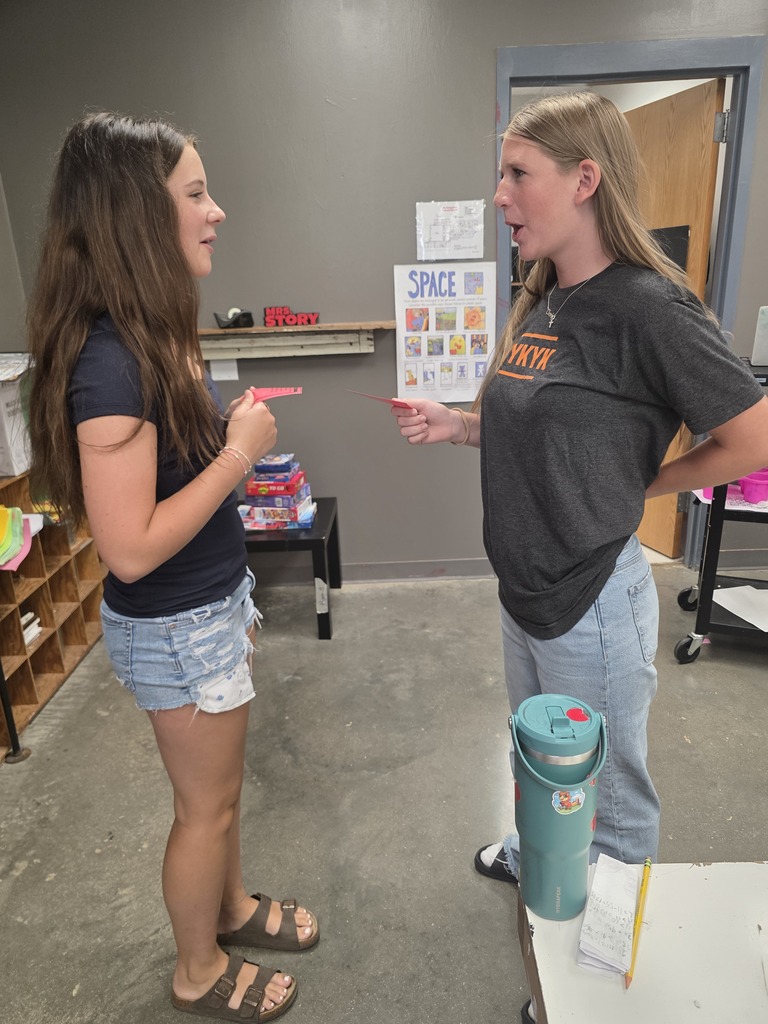 Two women standing, one gesturing with a finger, inside a room with a table and a poster.