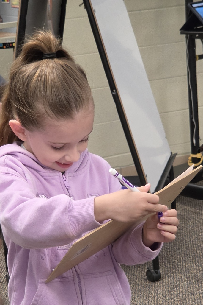 A young girl in a purple hoodie holds a clipboard and writes on it with a marker.