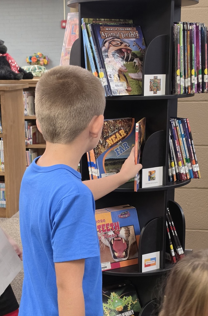 A boy in a blue shirt selects a book from a tall, black bookshelf. Another child, partially visible, stands nearby.
