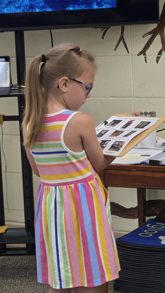 A young girl wearing a colorful dress and glasses stands at a table, examining a photo album.