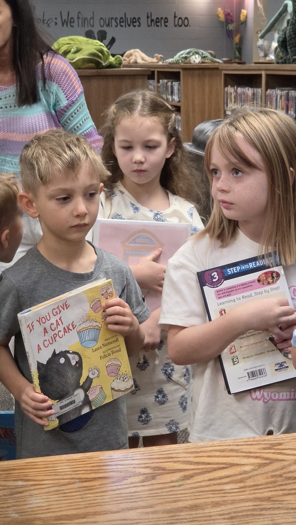Four children stand behind a counter holding books; two boys and two girls. The children are all holding books.
