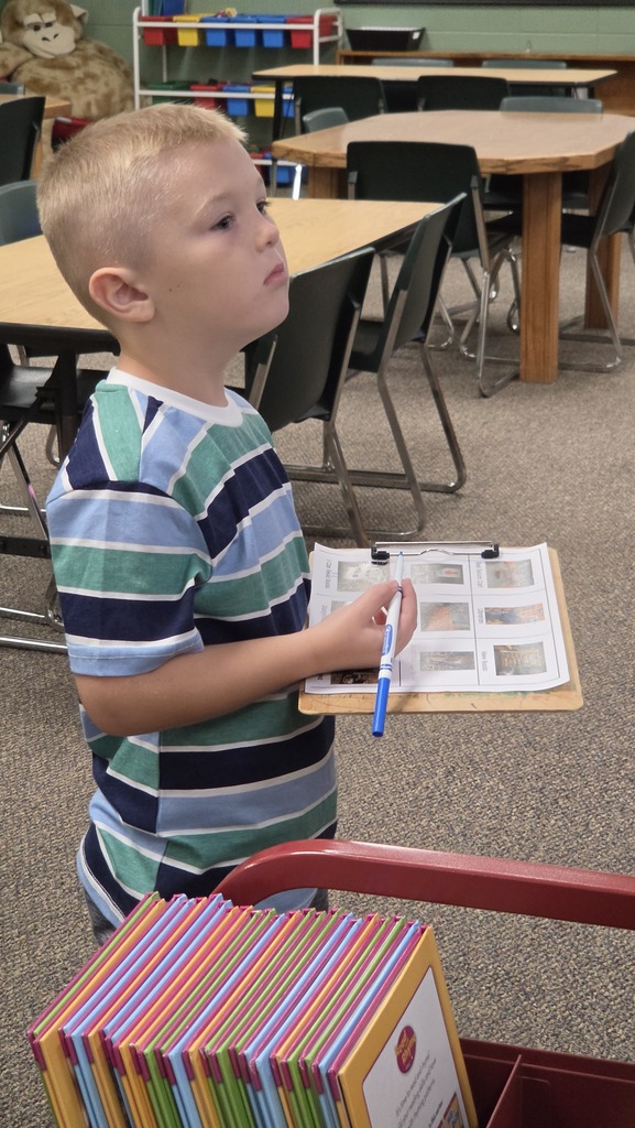 A child in a striped shirt holds a marker over a paper and stands next to a cart of books.