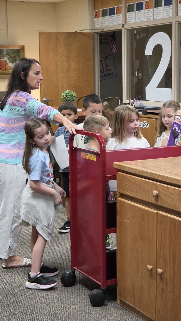 A woman in a striped shirt stands with children, showing them something. Behind them is a red cart and a wooden desk.