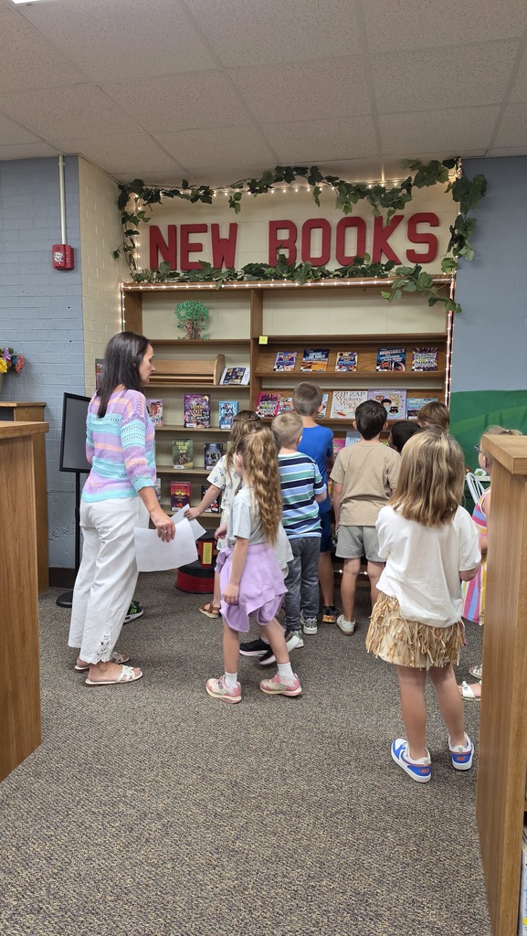 A woman stands with children in a library. A sign above reads "NEW BOOKS". Shelves filled with books are behind them.