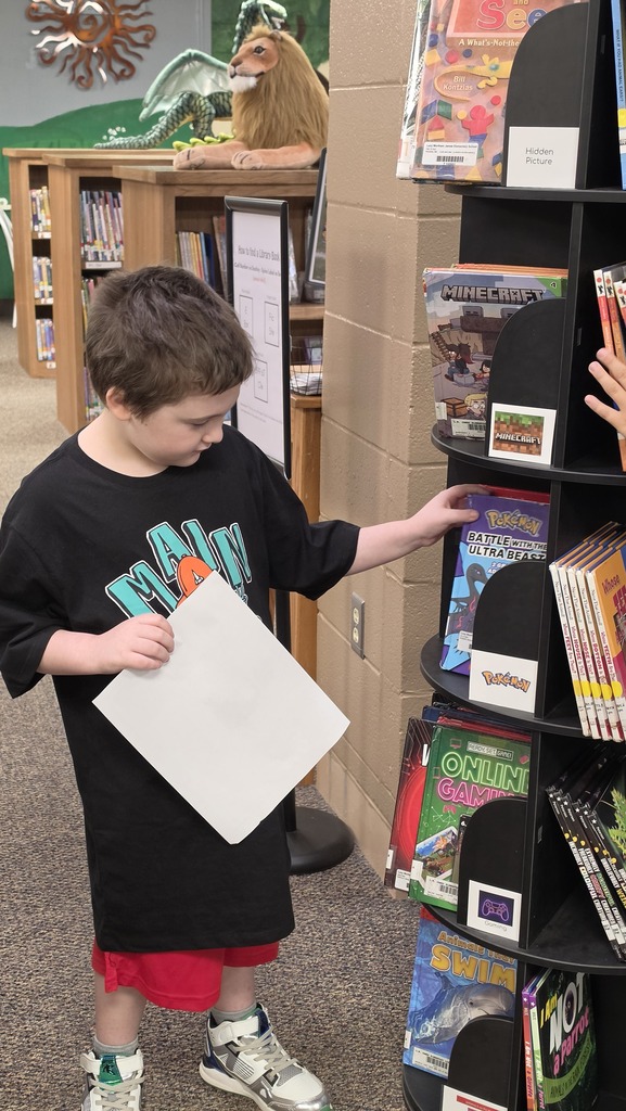 Boy in black t-shirt and red shorts stands in a library, holding a paper and reaching for books.