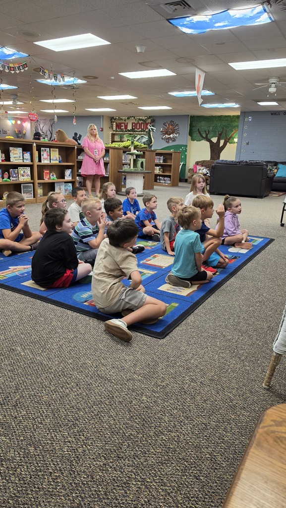 A group of children sits on a blue rug in a library, listening to a woman standing. Shelves and a tree-themed wall are in the background.