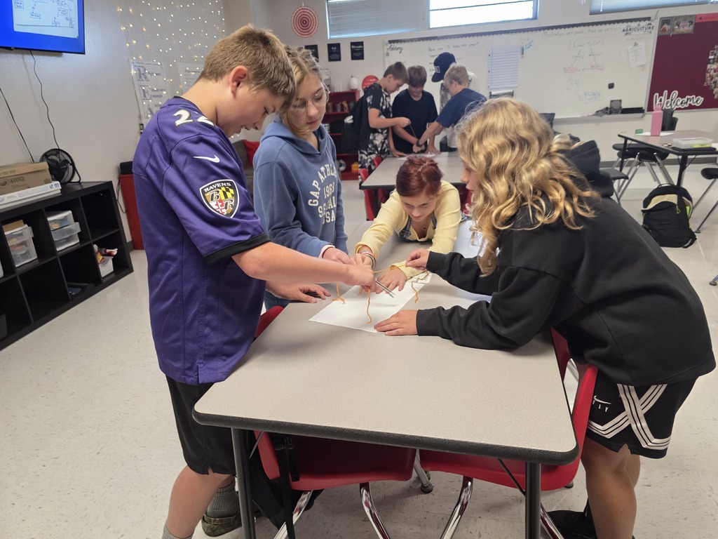 Team building in Mrs. Mulhall's class! Multiple students are working together to draw on a piece of paper with a single writing utensil and string. 