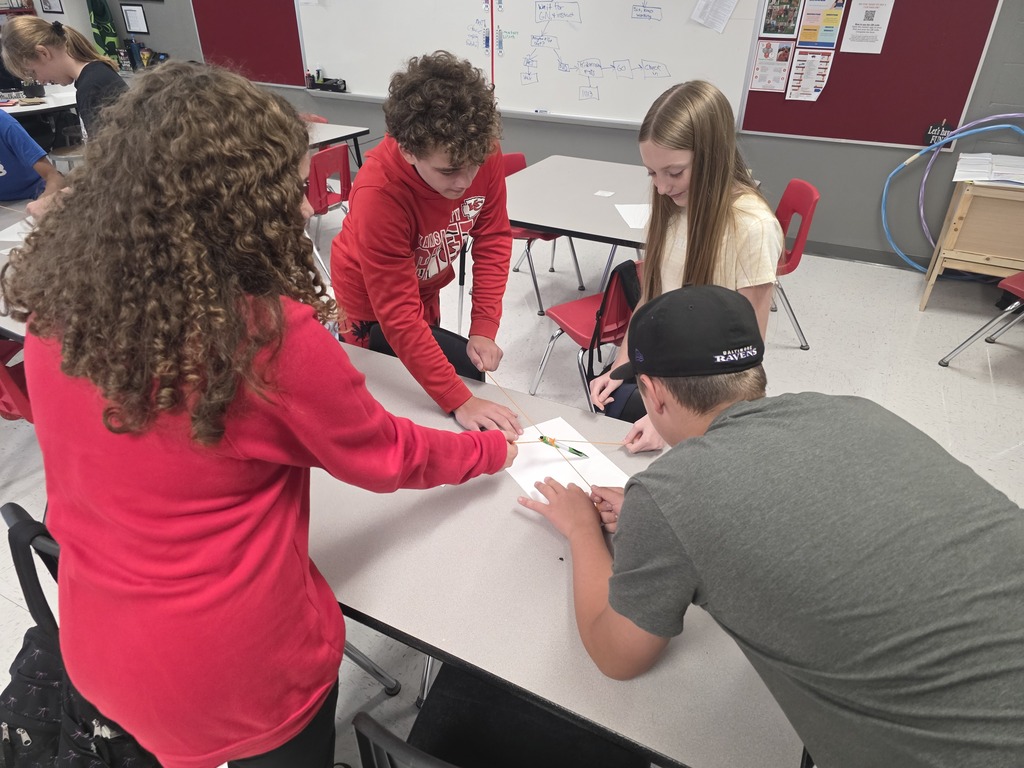 Team building in Mrs. Mulhall's class! Multiple students are working together to draw on a piece of paper with a single writing utensil and string. 