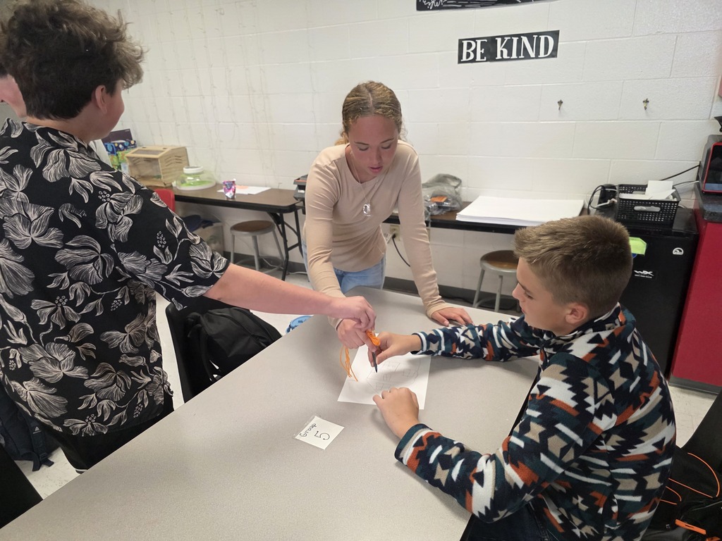 Team building in Mrs. Mulhall's class! Multiple students are working together to draw on a piece of paper with a single writing utensil and string. 
