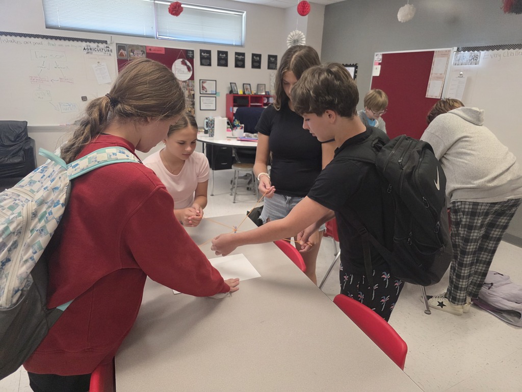 Team building in Mrs. Mulhall's class! Multiple students are working together to draw on a piece of paper with a single writing utensil and string. 