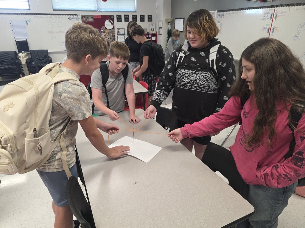 Team building in Mrs. Mulhall's class! Multiple students are working together to draw on a piece of paper with a single writing utensil and string. 
