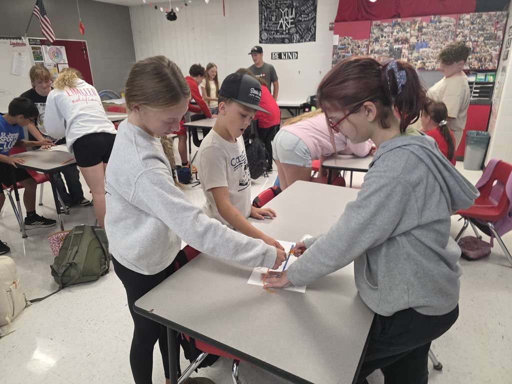 Team building in Mrs. Mulhall's class! Multiple students are working together to draw on a piece of paper with a single writing utensil and string. 