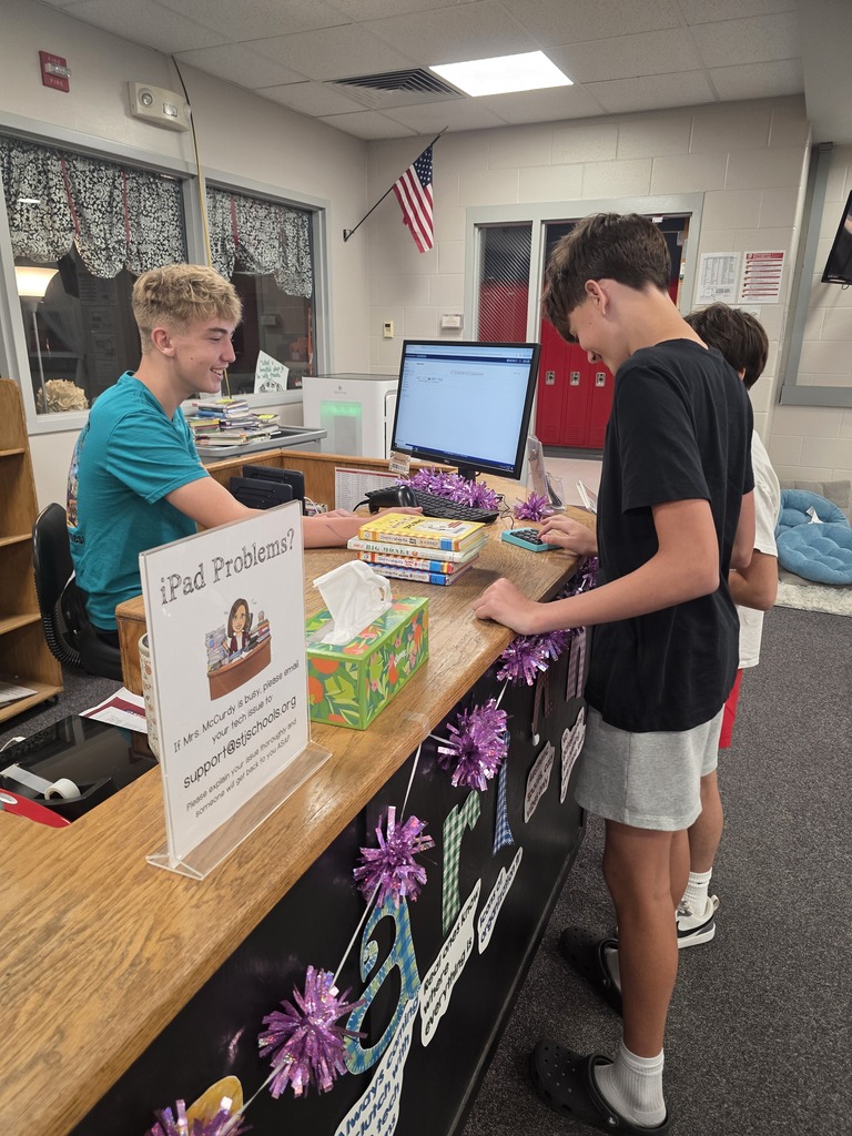 Two boys bend over a counter with books and decorations. One holds a book, the other points to a monitor. Behind, an American flag and windows.
