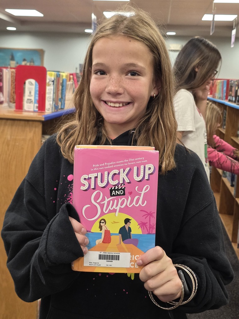 A girl in a library holds a book titled "Stuck Up" and "Stupid" while smiling.
