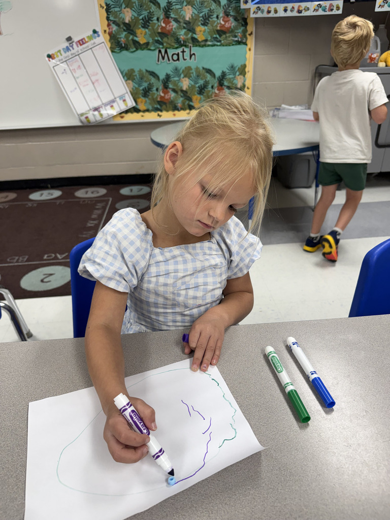 A young girl wearing a blue and white dress works on a drawing with colored markers.