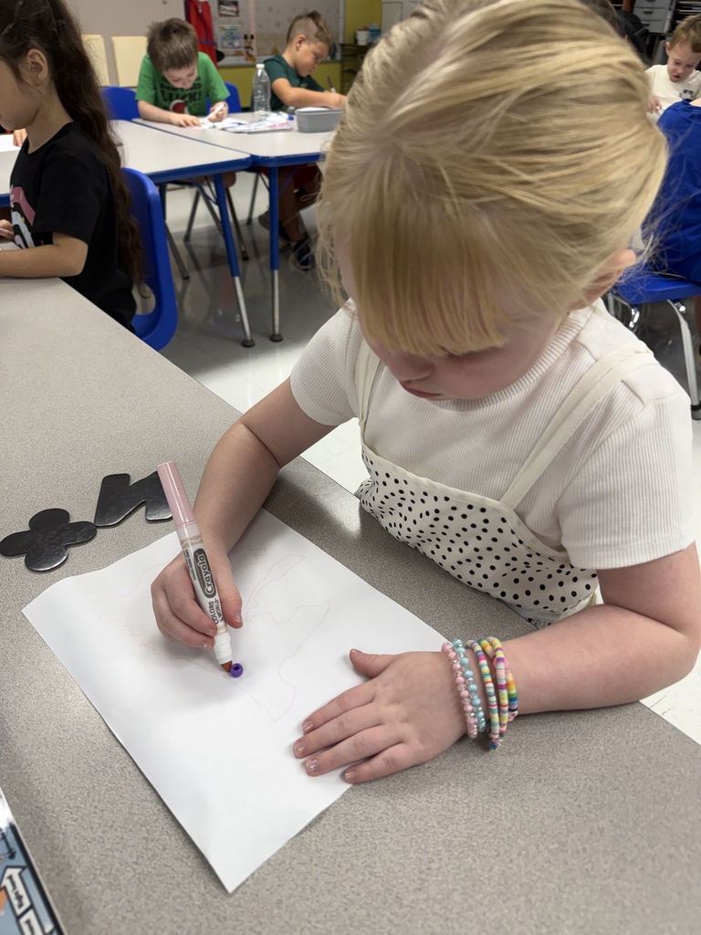Child focused on paper, holding a marker, surrounded by other students seated at tables in a classroom.