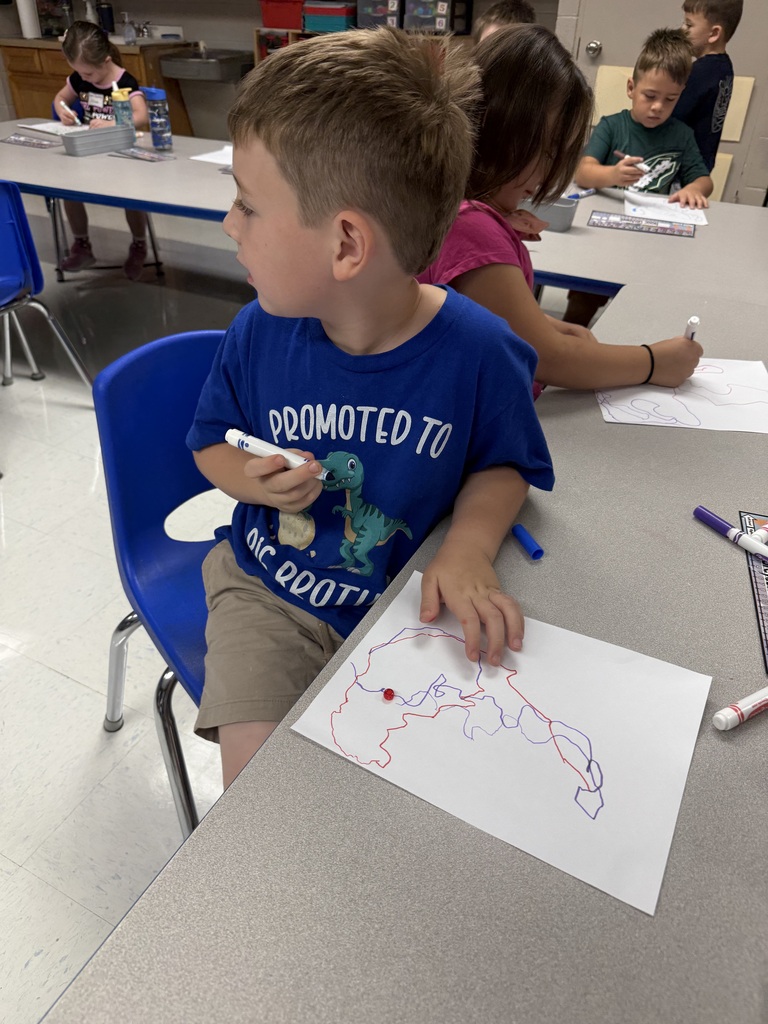 A young boy wearing a blue shirt sits in a chair and draws on paper.