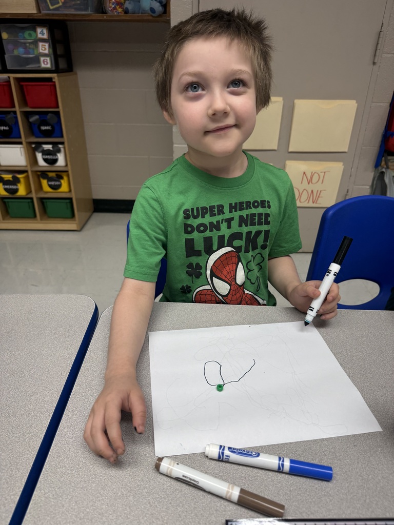 A child in a green t-shirt draws on a white sheet of paper with markers.