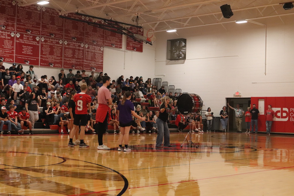 People gather on bleachers around a basketball court, with three individuals standing on the court. A photographer takes pictures.