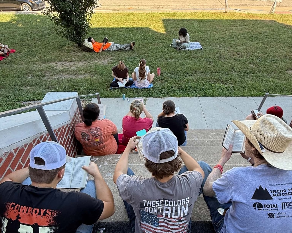 People sit on stairs and grass, some reading books, with a grassy background. Two wear cowboy hats.