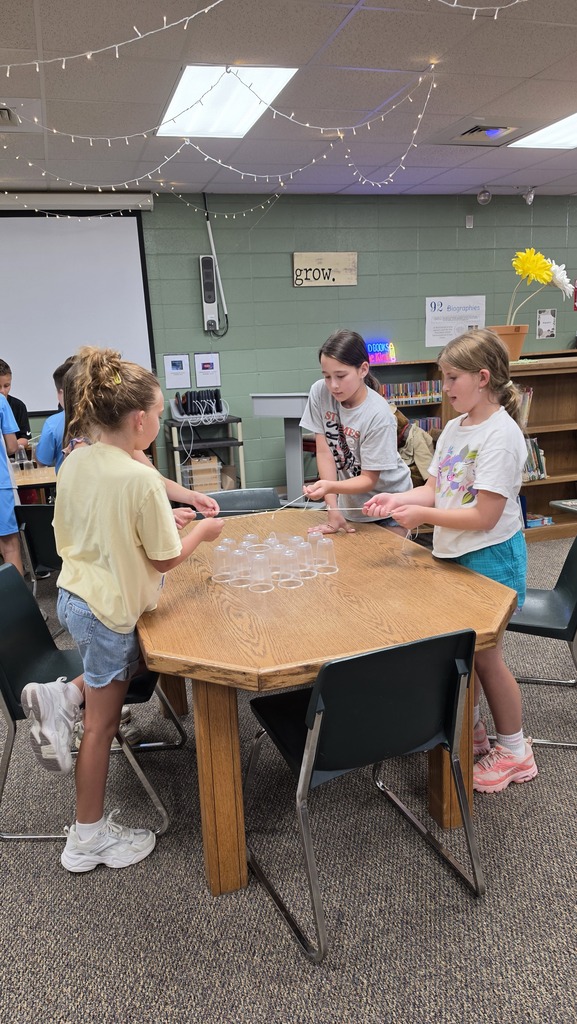 Four girls engage in an activity at a wooden octagon table in a classroom with bookshelves.