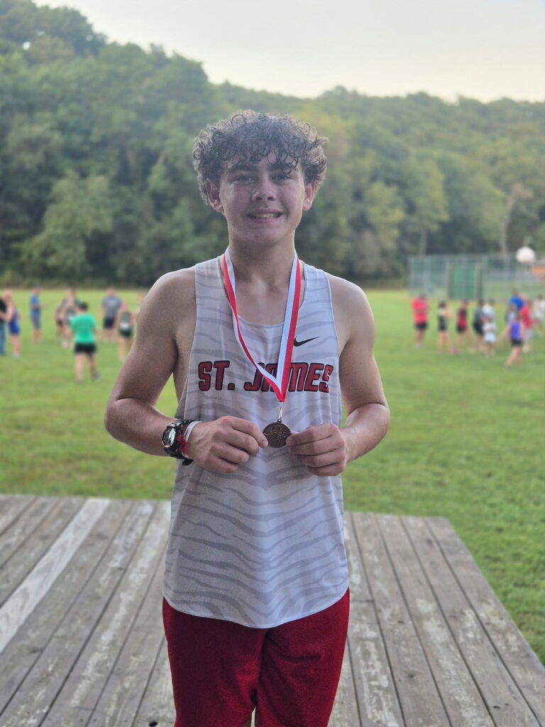 A smiling person in a gray tank top and red pants stands on a wooden platform. They hold two medals.
