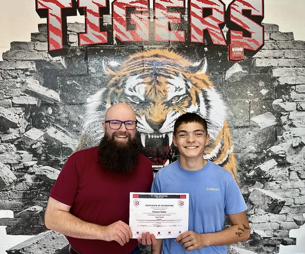 Two people stand in front of a tiger mural with a large word "Tigers". The man holds a certificate.